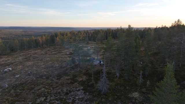 Ilmakuva avarasta, kumpuilevasta suomalaisesta metsämaisemasta Rovaniemen alueella tai Pohjois-Suomessa. Kuvassa näkyy sammaleen ja varvikon peittämää maastoa, harvakseltaan kasvavia mäntyjä ja kuusia. Keskellä, puiden lomassa, on pieni savuava nuotio ja sen äärellä ihminen istumassa tai seisomassa. Taustalla horisonttiin asti levittäytyy laaja metsäalue. Taivas on vaaleansininen ja vaaleanpunainen auringonlaskun aikaan, luoden kuvaan rauhallisen ja pohjoisen valon tunnelman.