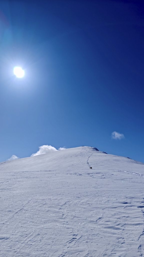 Pystysuuntainen kuva jyrkästä, puuttomasta ja lumipeitteisestä tunturin rinteestä. Rinteessä näkyy kaksi pientä, tummaa hahmoa (hiihtäjiä tai vaeltajia) nousemassa kohti huippua, jota kohti kulkee selkeä ura lumessa. Taivas on kirkkaan, syvän sininen, ja vasemmalla ylhäällä paistaa kirkas, valkoinen aurinko. Rinteen luminen pinta on epätasainen ja heijastaa auringonvaloa. Kuva luo voimakkaan tunteen haasteesta, korkeudesta ja kirkkaasta kevätauringosta Lapin tai Pohjois-Norjan tunturimaisemassa.