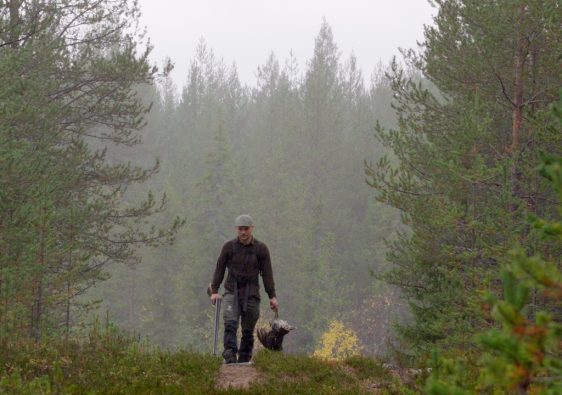 Mies kävelee eteenpäin sumuisessa metsämaastossa Pohjois-Suomessa. Hänellä on metsästysvaatteet ja -lakki, ja hän kantaa oikeassa kädessään saalista, luultavasti koppeloa tai muuta metsäkanalintua, ja vasemmassa kädessä ampuma-asettaan. Kuvaa kehystää tiheä havumetsä.