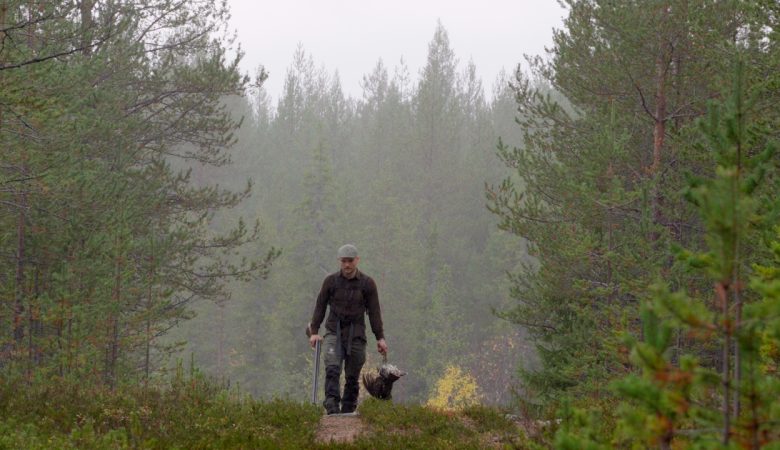 Mies kävelee eteenpäin sumuisessa metsämaastossa Pohjois-Suomessa. Hänellä on metsästysvaatteet ja -lakki, ja hän kantaa oikeassa kädessään saalista, luultavasti koppeloa tai muuta metsäkanalintua, ja vasemmassa kädessä ampuma-asettaan. Kuvaa kehystää tiheä havumetsä.