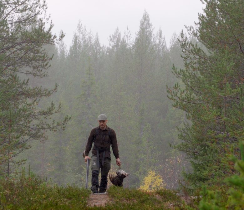 Mies kävelee eteenpäin sumuisessa metsämaastossa Pohjois-Suomessa. Hänellä on metsästysvaatteet ja -lakki, ja hän kantaa oikeassa kädessään saalista, luultavasti koppeloa tai muuta metsäkanalintua, ja vasemmassa kädessä ampuma-asettaan. Kuvaa kehystää tiheä havumetsä.