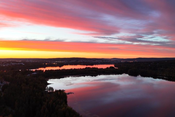 Ilmakuva rauhallisesta järvestä auringonlaskussa, jossa taivas hehkuu kirkkaissa pinkin, oranssin ja violetin sävyissä metsien ja kaukaisten mäkien yläpuolella.
