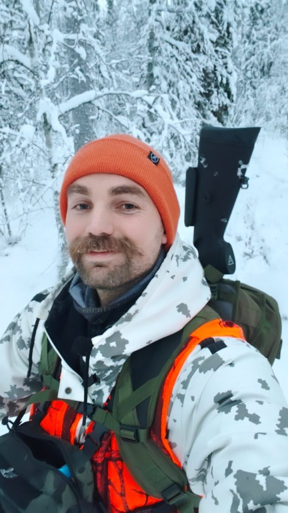 Lähikuva miehestä (Torsti/Kalle) talvisella hirvimetsällä. Hänellä on kirkkaan oranssi pipo, oransseja turvallisuusyksityiskohtia sisältävä valkoinen maastokuvioitu talvitakki ja reppu, jossa on kivääri. Taustalla on lumisateen peittämä metsä.