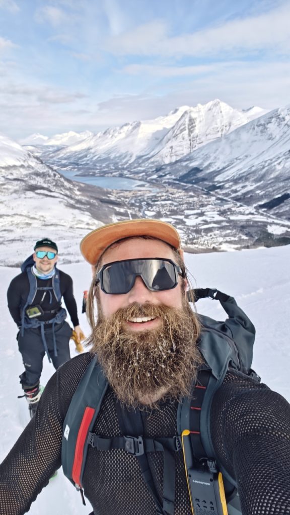Kaksi iloista miestä ottaa selfien korkealla lumisella vuorenrinteellä Lyngenissä, Norjassa. Kuvan etualalla oleva mies, jolla on paksu parta, oranssi lippalakki ja aurinkolasit, hymyilee leveästi. Hänen partaansa on kertynyt huurretta tai lunta. Taustalla näkyy toinen hiihtäjä/vaeltaja, joka hymyilee ja pitää kiinni repun olkaimesta. Heidän takanaan avautuu henkeäsalpaava vuoristomaisema, jossa lumiset huiput kehystävät alla olevaa sinistä merta tai järveä ja laaksoa. Kuva välittää onnistumisen, ilon ja ystävyyden tunteen jylhässä pohjoisessa ympäristössä.
