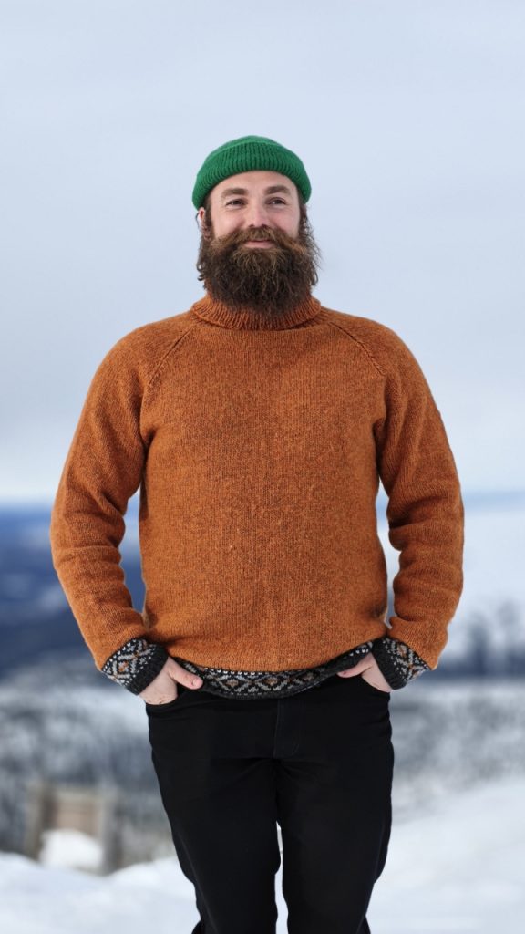 Hymyilevä mies lumisessa maisemassa, pukeutuneena oranssiin villapaitaan ja vihreään pipoon, kädet rentona taskuissa.