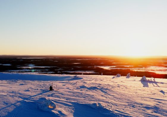 Laaja maisemakuva Luoston tai muun Lapin tunturin huipulta auringonlaskun aikaan. Etualalla on puuton, syvän lumen peittämä tunturialue, jonka sinertävät varjot korostavat lumen pinnan muotoja ja heijastavat auringonlaskun hehkua. Keskellä etualaa, hieman vasemmalla, näkyy yksinäinen hahmo istumassa tai kyyristelemässä. Taustalla horisontissa on laajoja, tummia metsäalueita ja järviä, joiden yllä auringonlasku värjää taivaan kirkkaan keltaisen, oranssin ja vaaleanpunaisen sävyillä. Tunnelma on avara, jylhä ja rauhallinen.
