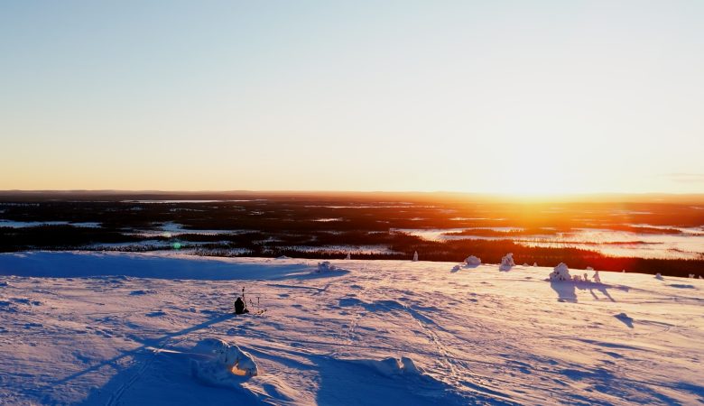 Laaja maisemakuva Luoston tai muun Lapin tunturin huipulta auringonlaskun aikaan. Etualalla on puuton, syvän lumen peittämä tunturialue, jonka sinertävät varjot korostavat lumen pinnan muotoja ja heijastavat auringonlaskun hehkua. Keskellä etualaa, hieman vasemmalla, näkyy yksinäinen hahmo istumassa tai kyyristelemässä. Taustalla horisontissa on laajoja, tummia metsäalueita ja järviä, joiden yllä auringonlasku värjää taivaan kirkkaan keltaisen, oranssin ja vaaleanpunaisen sävyillä. Tunnelma on avara, jylhä ja rauhallinen.