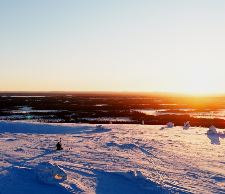 Laaja maisemakuva Luoston tai muun Lapin tunturin huipulta auringonlaskun aikaan. Etualalla on puuton, syvän lumen peittämä tunturialue, jonka sinertävät varjot korostavat lumen pinnan muotoja ja heijastavat auringonlaskun hehkua. Keskellä etualaa, hieman vasemmalla, näkyy yksinäinen hahmo istumassa tai kyyristelemässä. Taustalla horisontissa on laajoja, tummia metsäalueita ja järviä, joiden yllä auringonlasku värjää taivaan kirkkaan keltaisen, oranssin ja vaaleanpunaisen sävyillä. Tunnelma on avara, jylhä ja rauhallinen.