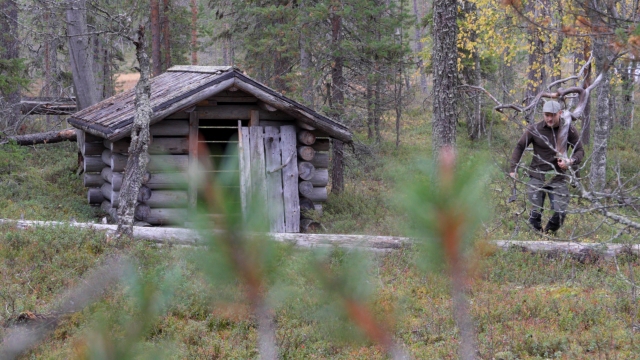Kuva vanhasta, hirsirakenteisesta laavusta metsässä. Hökkelin ovi on raollaan. Kuvan oikealla puolella ja taka-alalla näkyy metsämaastossa juokseva tai kävelevä mies, jolla on metsästysvaatteet ja lippalakki.