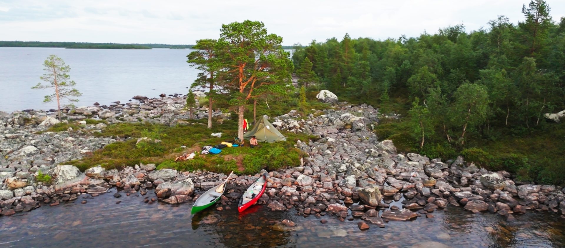Ilmakuva kahdesta rannalla kelluvasta kanootista (vihreä ja punainen) kivisellä rannalla Vätsärin erämaa-alueella. Rannalla on kiveyden ja pienten puiden keskellä teltta ja leirivarusteita. Taustalla näkyy laaja järvi ja tiheää metsää.